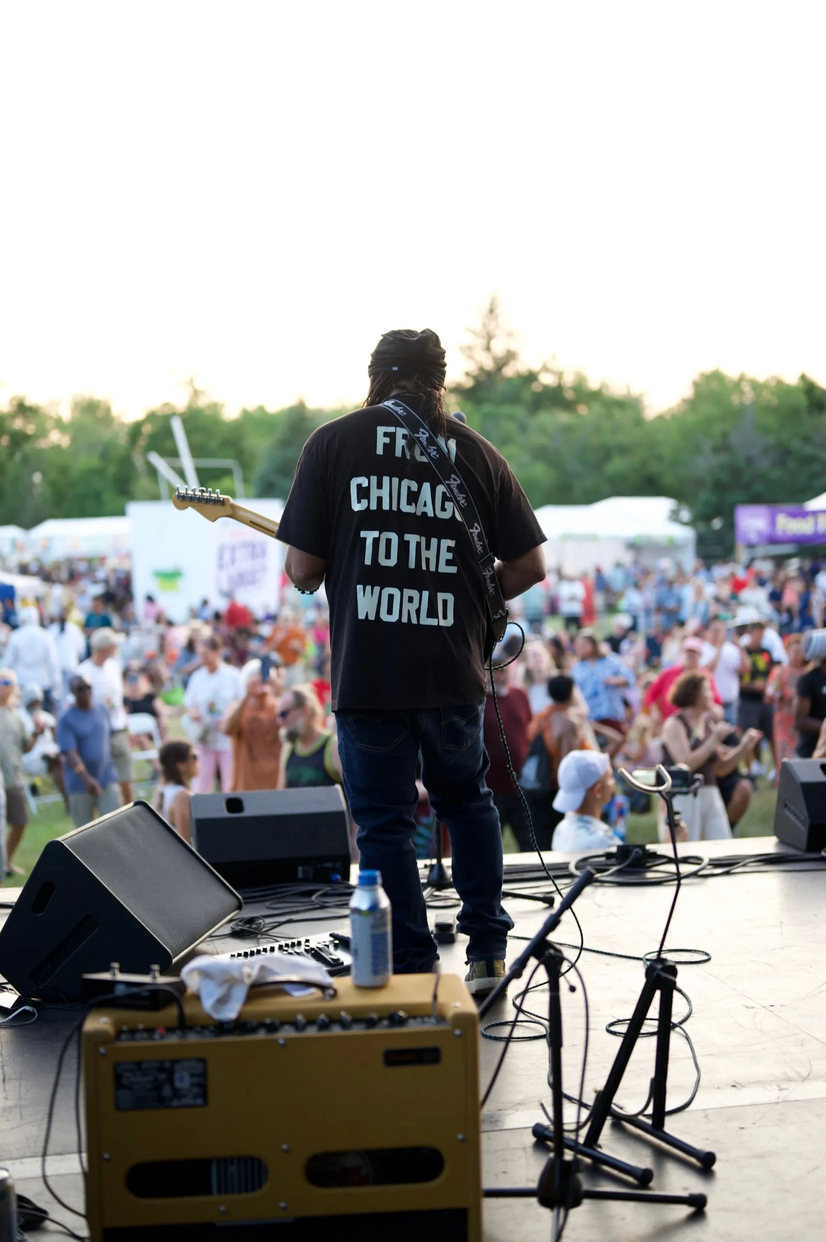 A performer seen from behind, wearing a "From Chicago to the World" shirt, facing a festival crowd