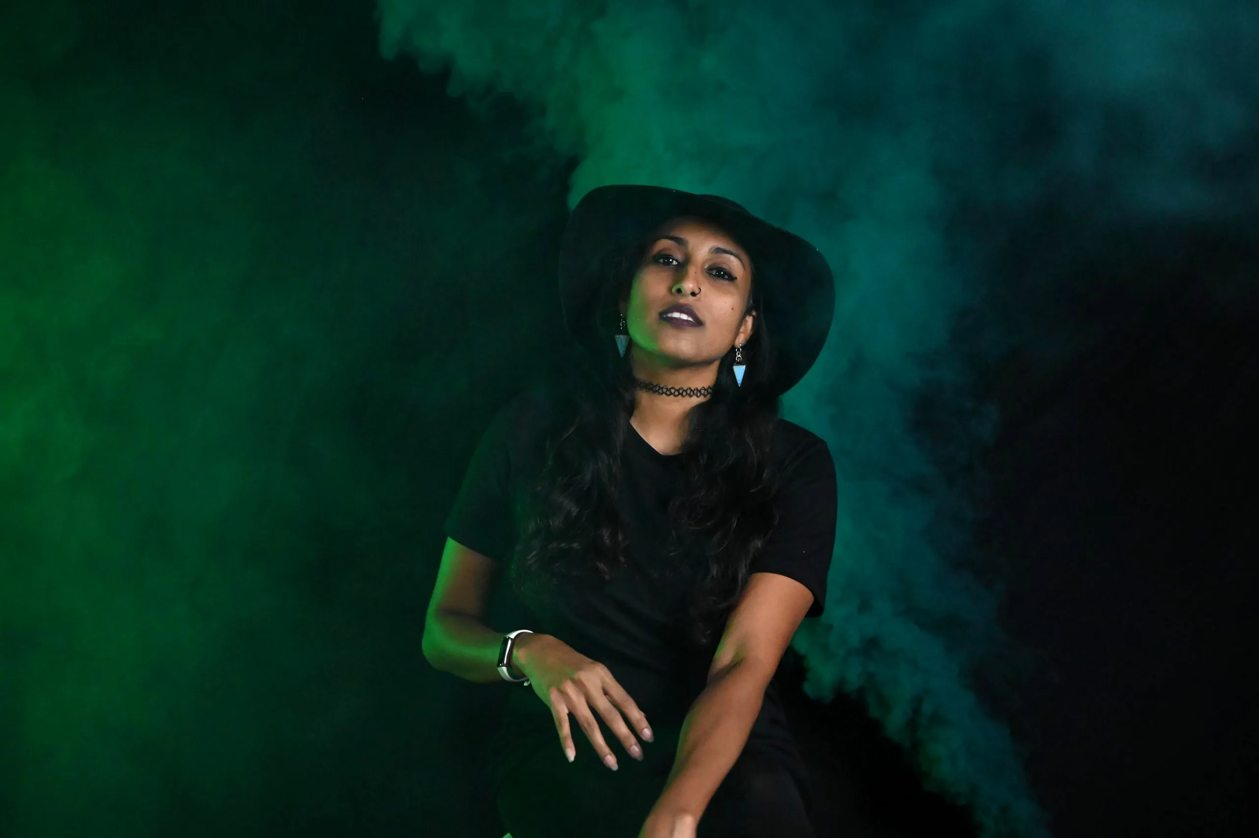 A woman in a black wide-brimmed hat and black dress, seated in swirling green smoke